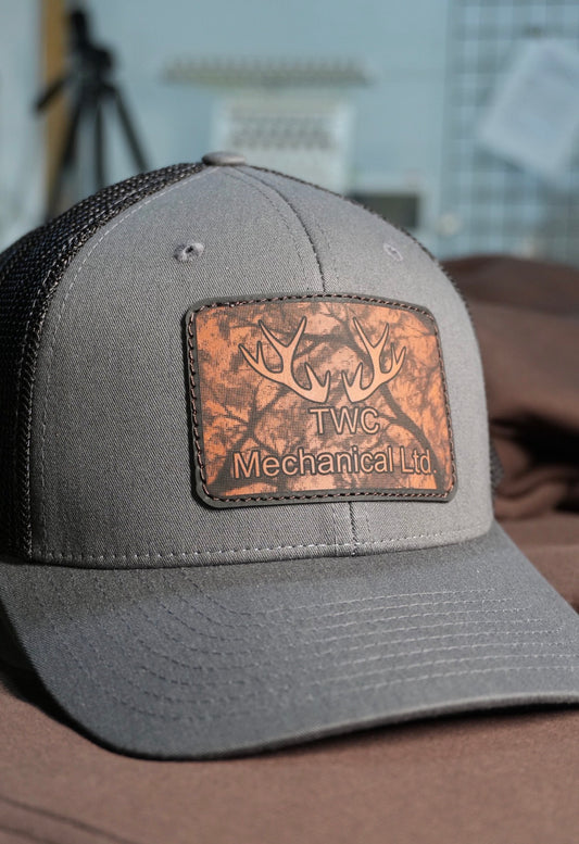 Gray cap with a leather patch featuring 'TWC Mechanical Ltd' on a blurred background